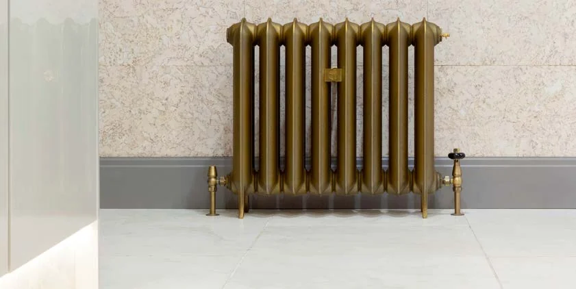 Castrads Neptune cast iron radiator in a brass finish against a stone-tiled bathroom wall